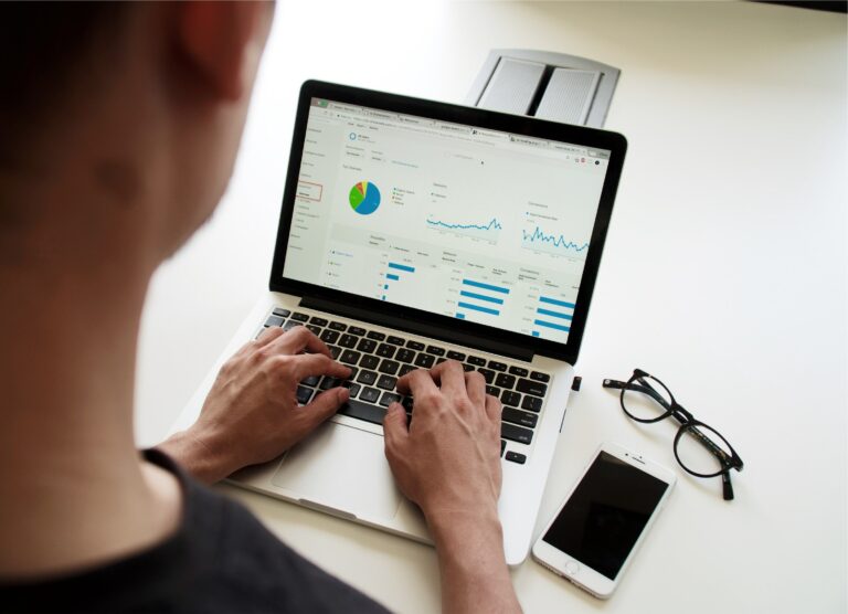 An accountant working on a laptop with charts and reports on the screen and glasses and phone on the table to automate the accounting services management process